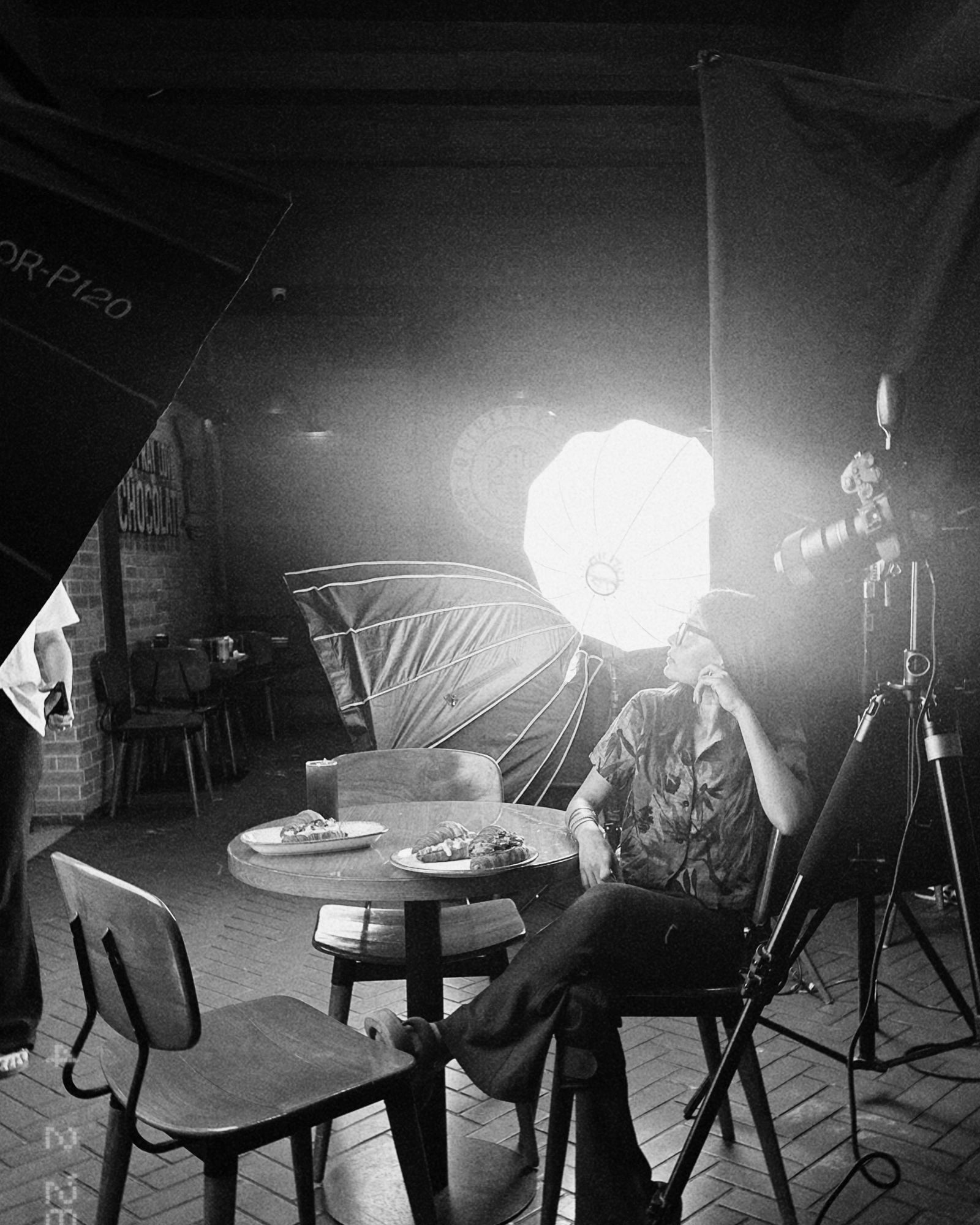 Nandika Shah on set, seated beside a café table of plated croissants, looking up into a lit softbox with her camera on a tripod beside her